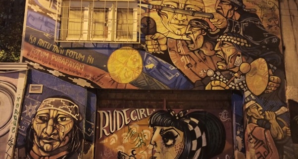 Wandbild in La Paz, Bolivien, aufgenommen am 8. April 2025: Die farbenfrohe Streetart zeigt indigene Figuren in traditioneller Kleidung und kämpferischen Posen, begleitet von Symbolen wie Trommeln, Speeren und goldenen Sonnen. Links ist ein Porträt einer ernsten Person mit traditionellem Stirnband zu sehen. In der Mitte prangt ein Text auf Spanisch und Mapudungun sowie ein Graffiti mit dem Schriftzug 'RUDEGIRL' und 'Stay Rebel'.